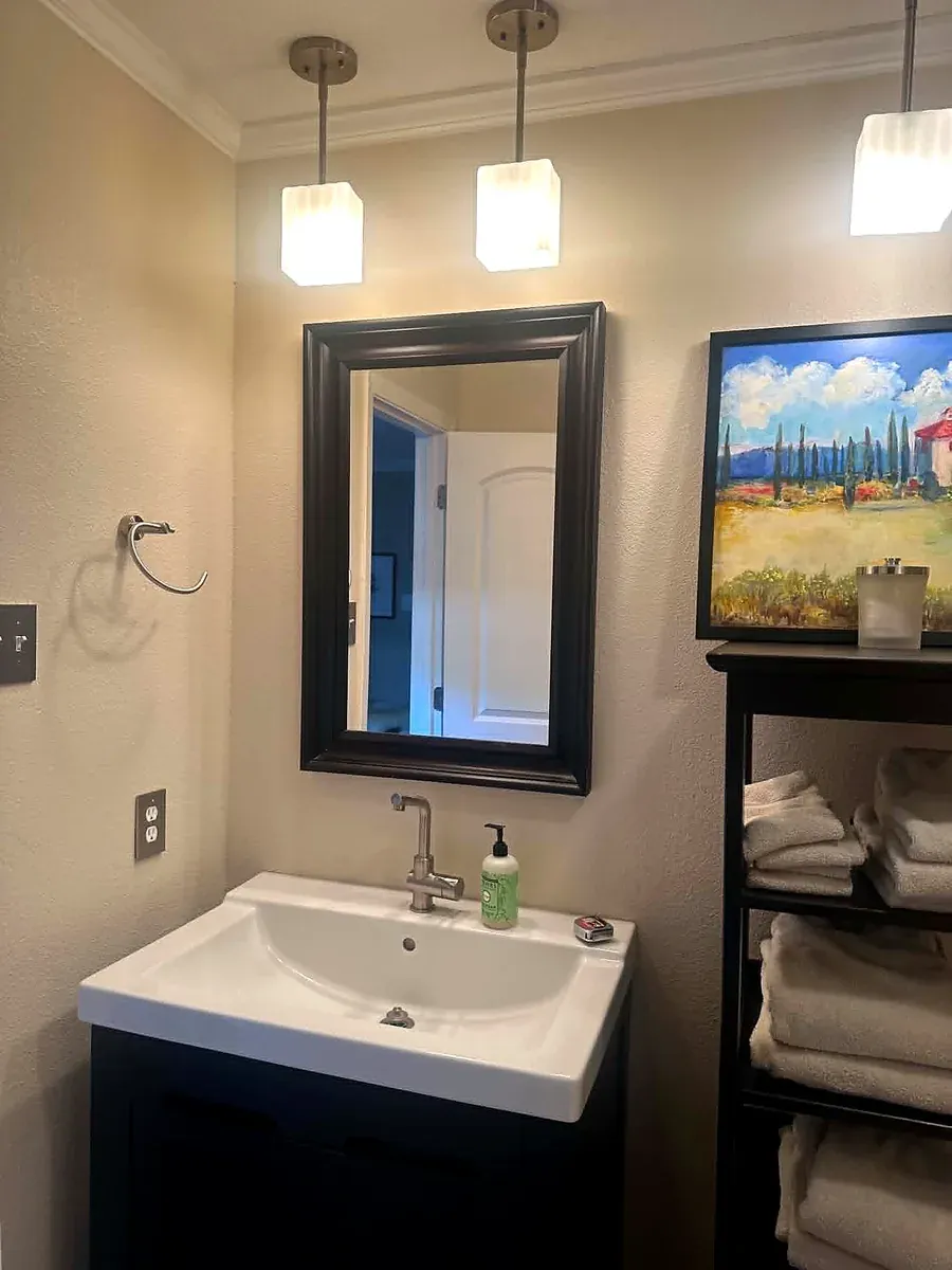Powder bathroom with a dark vanity, framed mirror, wall sconce and a floating shelf tower
